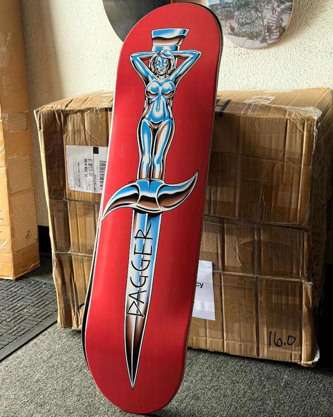 Dagger Chrome Angel skateboard deck standing upright, featuring Woogie’s silver dagger-and-figure artwork on a red deck.